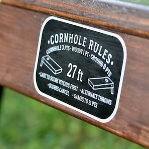 Puede incluir: Pegatina en blanco y negro con el texto "&bull;CORNHOLE RULES." y "27 ft" con instrucciones para jugar al cornhole. La pegatina est&aacute; en una superficie de madera marr&oacute;n.