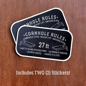 Puede incluir: Dos pegatinas en blanco y negro con el texto "Cornhole Rules" e instrucciones para jugar al juego. Las pegatinas est&aacute;n separadas 27 pies. El texto en las pegatinas dice: "Cornhole 3 pts - Woody 1 pt - Ground 0 pts" y "Last to score pitches first - Alternate throws - Scores cancel - Games to 21 pts".