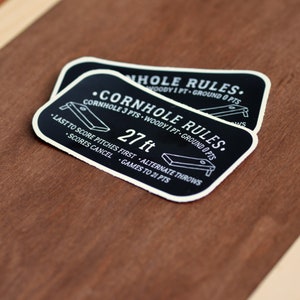 Puede incluir: Pegatina en blanco y negro con el texto "CORNHOLE RULES." y un diagrama de un tablero de cornhole. El texto tambi&eacute;n incluye las reglas: "CORNHOLE 3 PTS - WOODY 1 PT - GROUND 0 PTS", "LAST TO SCORE PITCHES FIRST - ALTERNATE THROWS", "SCORES CANCEL - GAMES TO 21 PTS", y "27 ft".