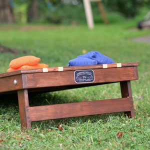Puede incluir: Un juego de cornhole de madera con dos sacos de frijoles, uno naranja y uno azul. El tablero del juego tiene un letrero negro que dice "Cornhole Rules 27 ft."