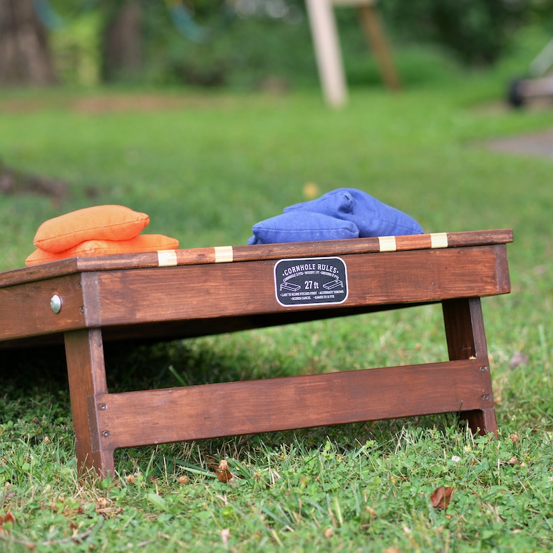 Puede incluir: Un juego de cornhole de madera con dos sacos de frijoles, uno naranja y uno azul. El tablero del juego tiene un letrero negro que dice "Cornhole Rules 27 ft."