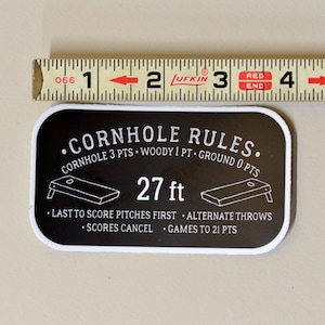 Puede incluir: Pegatina en blanco y negro con el texto "CORNHOLE RULES." y las reglas del juego. La pegatina incluye un diagrama de un tablero de cornhole y el texto "27 ft".