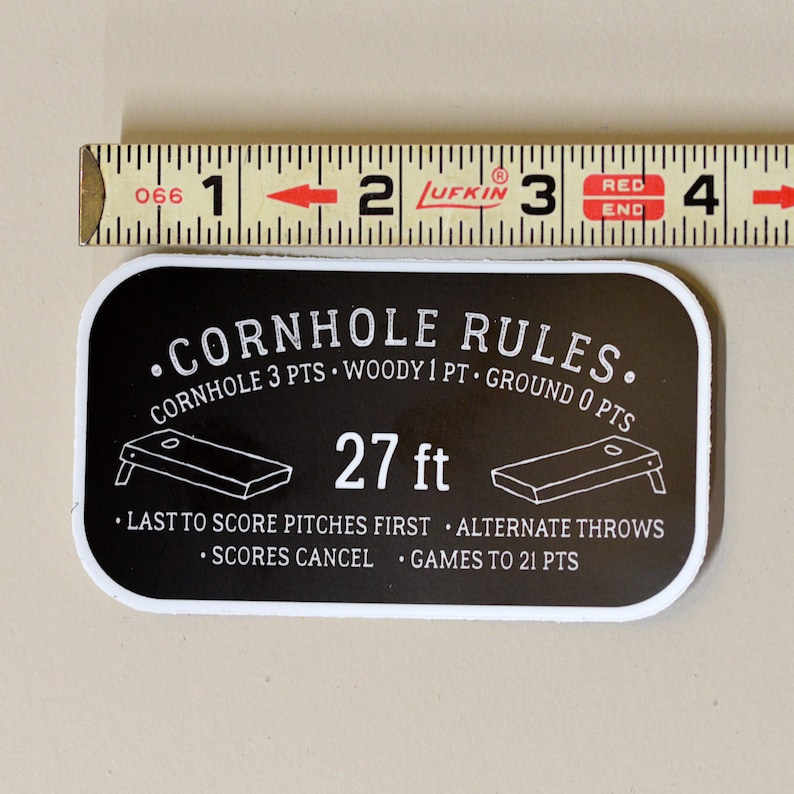 Puede incluir: Pegatina en blanco y negro con el texto "CORNHOLE RULES." y las reglas del juego. La pegatina incluye un diagrama de un tablero de cornhole y el texto "27 ft".