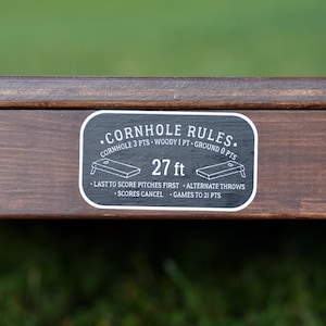 Puede incluir: Una pegatina en blanco y negro con el texto "&bull;CORNHOLE RULES." y "CORNHOLE 3 PTS &bull; WOODY 1 PT &bull; GROUND O PTS" y "27 ft". La pegatina tambi&eacute;n incluye instrucciones para jugar al cornhole, como "LAST TO SCORE PITCHES FIRST &bull; ALTERNATE THROWS &bull; SCORES CANCEL &bull; GAMES TO 21 PTS".