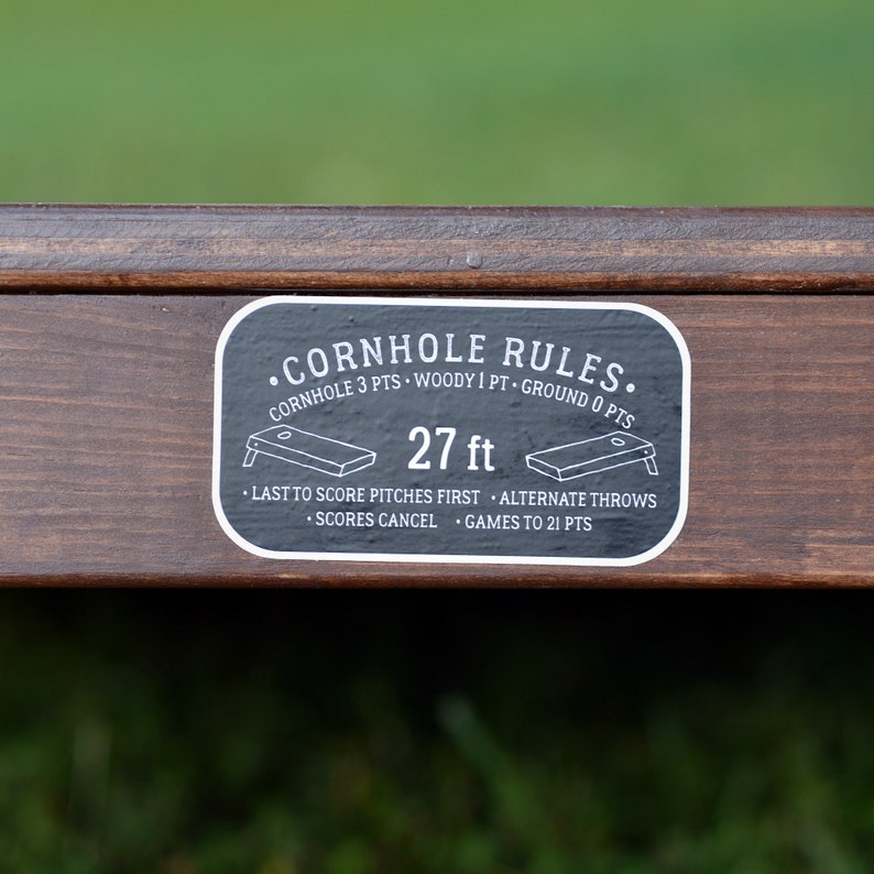 Puede incluir: Una pegatina en blanco y negro con el texto "&bull;CORNHOLE RULES." y "CORNHOLE 3 PTS &bull; WOODY 1 PT &bull; GROUND O PTS" y "27 ft". La pegatina tambi&eacute;n incluye instrucciones para jugar al cornhole, como "LAST TO SCORE PITCHES FIRST &bull; ALTERNATE THROWS &bull; SCORES CANCEL &bull; GAMES TO 21 PTS".