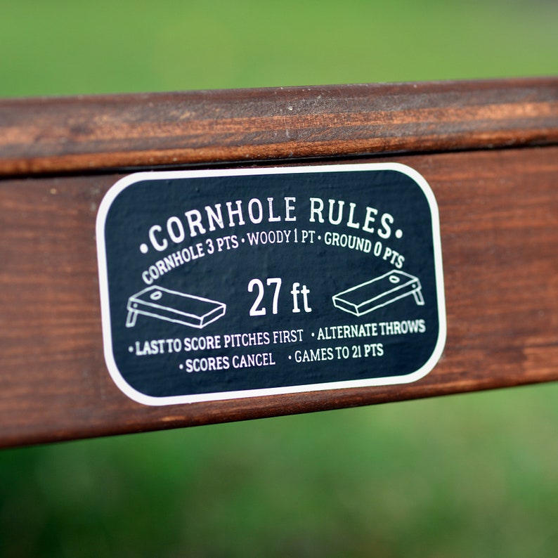 Puede incluir: Pegatina en blanco y negro con el texto "CORNHOLE RULES." e instrucciones para jugar al cornhole. La pegatina est&aacute; sobre una superficie de madera marr&oacute;n. El texto de la pegatina dice: "CORNHOLE 3 PTS. WOODY 1 PT. GROUND 0 PTS. 27 ft. LAST TO SCORE PITCHES FIRST. ALTERNATE THROWS. SCORES CANCEL. GAMES TO 21 PTS."