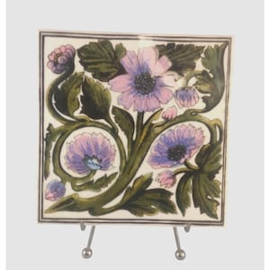 May include: Square ceramic tile with a floral design. The tile features lavender flowers with dark centers, green leaves, and buds against a cream background. The tile is displayed on a silver metal stand.