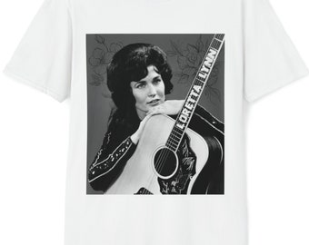 Può includere: Una maglietta bianca con una foto in bianco e nero di Loretta Lynn che tiene una chitarra. La chitarra ha la scritta "Loretta Lynn".