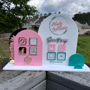 May include: A white and pink acrylic display with social media icons and QR codes. The display includes the text "Freaky by Kadin" and "Scan to pay". A teal-colored stand is attached to the base. The display is on a wooden surface.
