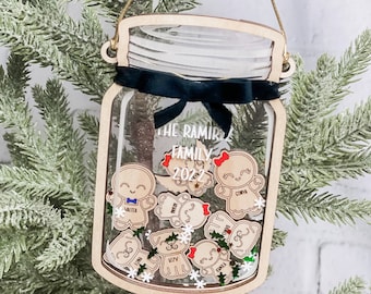 Puede incluir: Un adorno navideño de madera con forma de tarro de cristal con una cinta negra alrededor de la tapa. El tarro está lleno de figuras de pan de jengibre con nombres escritos en ellas, incluyendo Walter, Ivy y Emma. El tarro también tiene purpurina y copos de nieve en su interior. El texto "The Ramirez Family 2022" está escrito en el tarro.