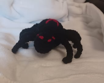 May include: A black crocheted spider with red eyes. The spider is lying on a white surface.