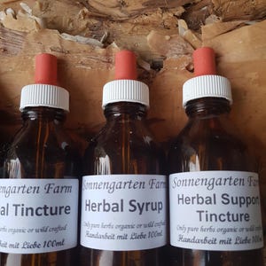 May include: Three brown glass bottles with white labels and red droppers. The labels read "Herbal Tincture", "Herbal Syrup", and "Herbal Support Tincture". The bottles are filled with a liquid and are against a textured, light brown background.
