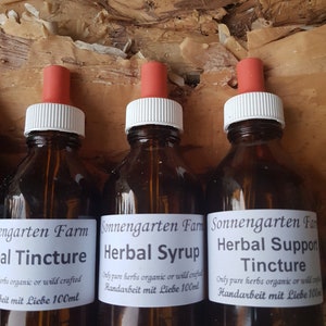 Op de afbeelding: Drie amberkleurige glazen flessen met druppelaars, gelabeld "Sonnengarten Farm" en gevuld met kruidentincturen en siropen. De etiketten zijn in zwarte tekst en luiden "Herbal Support Tincture", "Herbal Syrup" en "al Tincture". Elke fles bevat 100 ml.