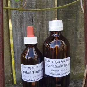 May include: Two brown glass bottles of herbal tincture with white labels and caps. The labels read "Sonnengarten Farm Herbal Tincture" and "Organic Herbal Tincture Concentrate 1:1". One bottle has a dropper. The background is a wooden fence.
