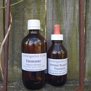 May include: Two glass bottles with dropper tops filled with brown liquid. The bottles have white labels with black text that reads "Sonnengarten Farm Immune Support Tincture Only pure herbs organic or wild crafted Handarbeit mit Liebe" and "Sonnengarten Farm Herbal Support Tincture Only pure herbs organic or wild crafted Handarbeit mit Liebe".