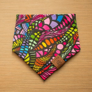 May include: A colorful dog bandana with a black background and a geometric pattern in shades of pink, orange, yellow, green, blue, and purple. The bandana has a brown leather tag with the word "Cedar" and a small flower design.