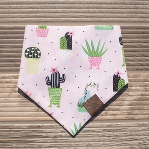 May include: Pink bandana with a cactus print. The bandana features a variety of cacti in different pots with black, green, and white details. The bandana has a brown leather patch on the bottom.