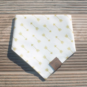 May include: White bandana with a gold arrow pattern. The bandana has a brown leather tag with the word "Pawsitive" on it.