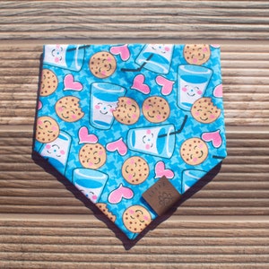 May include: A blue bandana with a pattern of cookies, glasses of milk, and hearts. The bandana has a brown leather tag with the word "CUPPA" on it.