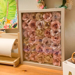 May include: A framed wall art piece featuring a collection of paper flowers in shades of pink and beige. The flowers are arranged in a grid pattern and are displayed against a white background.