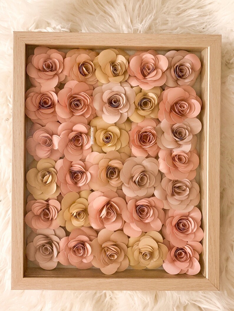 May include: A framed wall art piece featuring a collection of 25 paper flowers in shades of pink and beige. The flowers are arranged in a grid pattern and are all the same size and shape.