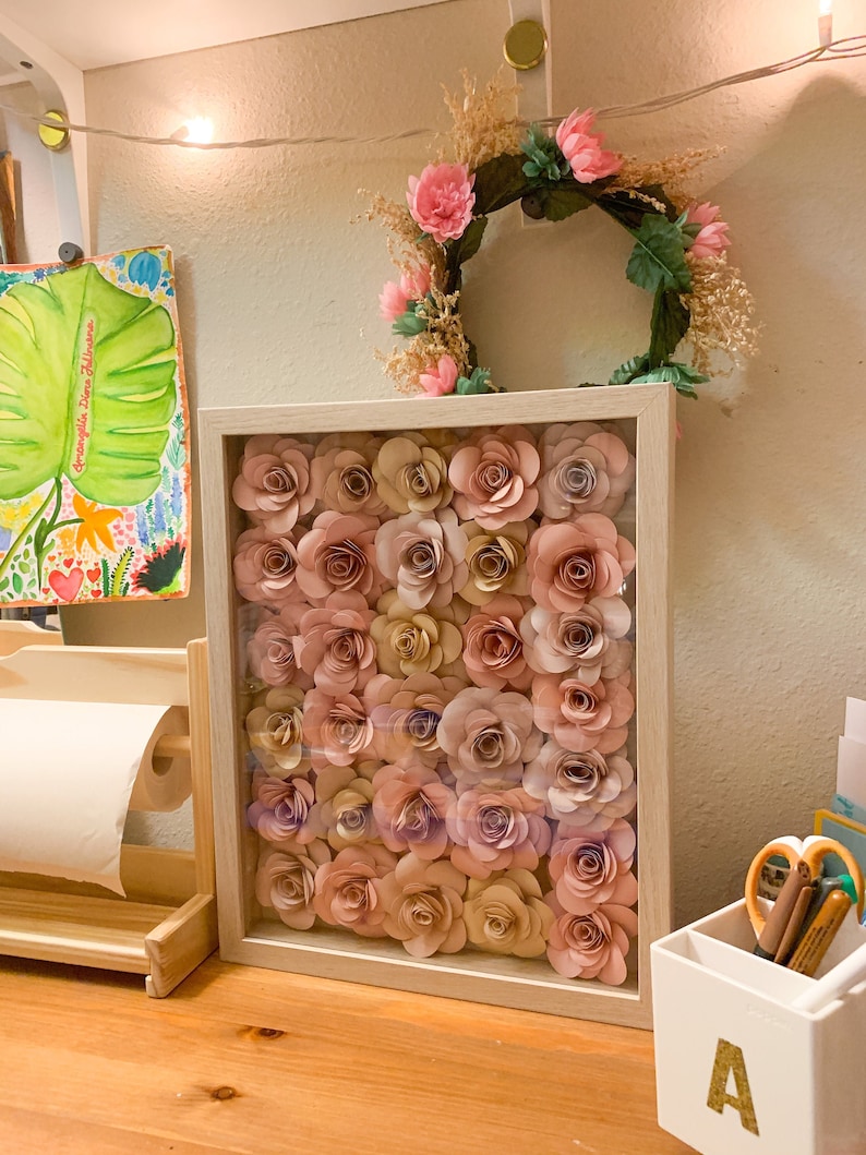 May include: A framed wall art piece featuring a collection of paper flowers in shades of pink and beige. The flowers are arranged in a grid pattern and are displayed against a white background.