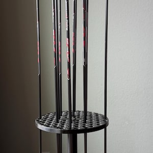 May include: A black arrow stand holding multiple arrows. The arrows have green and black fletching, red and yellow tips. The stand is made of black plastic and has a circular design with multiple holes.