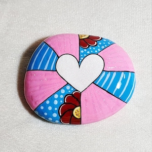 May include: A hand-painted, oval-shaped stone with a white heart in the center. The stone is divided into sections of pink, blue with white dots, and blue with white stripes. Red flowers with yellow centers are also painted on the stone.