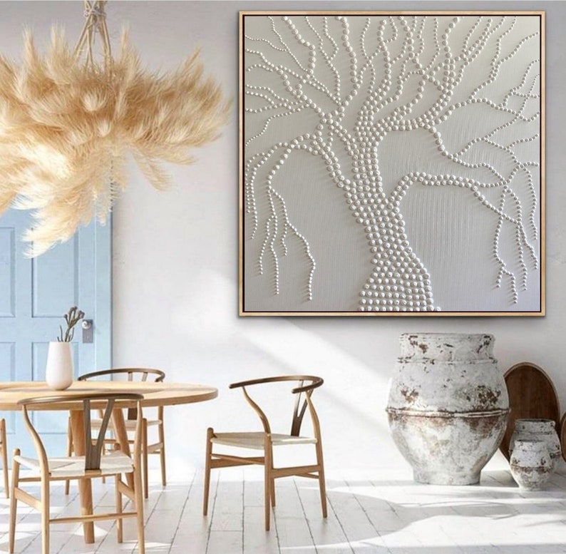 May include: A white canvas painting with a textured tree design made of white beads. The tree is centered on the canvas and has a thick trunk and branches that extend to the edges of the canvas. The painting is framed in a thin, gold frame.