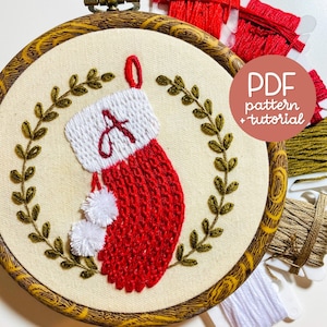 Op de afbeelding: Geborduurde kerstsok met de letter A in wit en rood garen, omringd door een groene krans op een beige achtergrond. De sok bevindt zich in een houten borduurring. De tekst "PDF-patroon + tutorial" staat op een roze achtergrond.