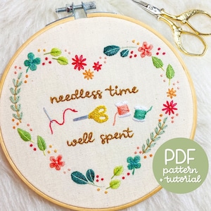 Peut inclure: Art de broderie sur un cercle avec une couronne en forme de cœur de feuilles vertes et de fleurs colorées. Le texte "needless time well spent" est brodé au centre, ainsi qu'une aiguille, du fil et des ciseaux. Le cercle est en bois et a un fond en tissu blanc. Le texte "PDF pattern + tutorial" est écrit en vert sur un cercle blanc dans le coin inférieur droit.