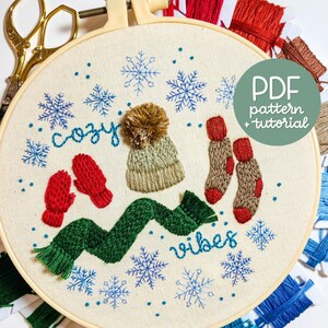 Peut inclure: Art de broderie sur un cercle avec une écharpe verte, des mitaines rouges, un bonnet en tricot beige et marron avec un pompon marron, et deux chaussettes marron et rouges. L'art du cercle comprend également les mots "cozy vibes" et des flocons de neige bleus. Le cercle est entouré de fil à broder.