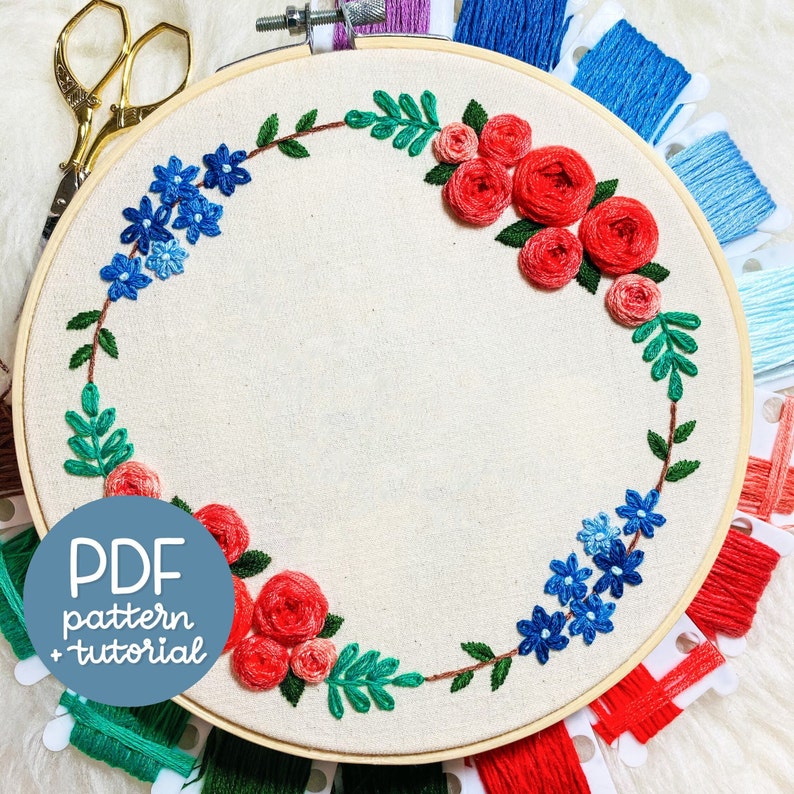 May include: A white embroidery hoop with a floral design. The design features red roses, blue flowers, and green leaves. The text "PDF pattern + tutorial" is printed on the hoop.