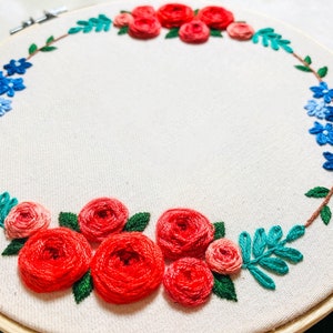 May include: A circular embroidery hoop with a white fabric background. The fabric is embroidered with a wreath of red and pink roses, blue flowers, and green leaves.