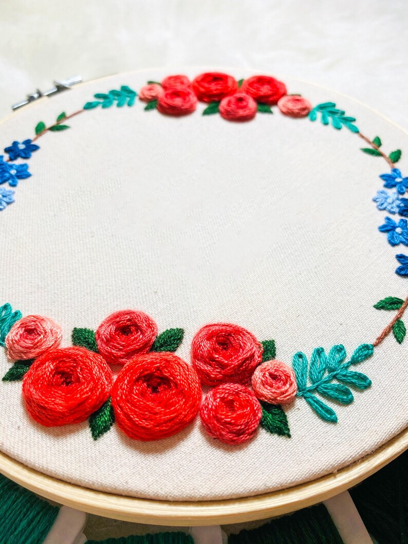 May include: A circular embroidery hoop with a white fabric background. The fabric is embroidered with a wreath of red and pink roses, blue flowers, and green leaves.
