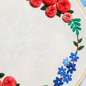 May include: A white embroidery hoop with a floral design. The design features red roses and blue flowers with green leaves. The hoop is made of wood and has a natural finish.