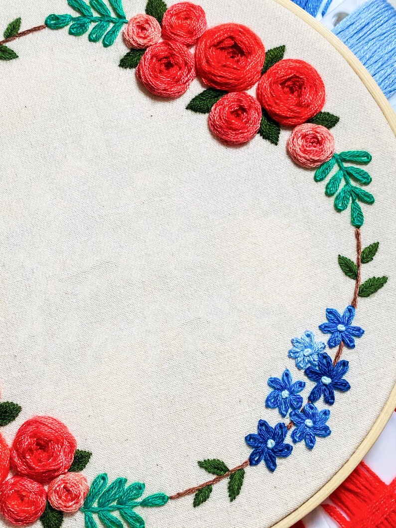 May include: A white embroidery hoop with a floral design. The design features red roses and blue flowers with green leaves. The hoop is made of wood and has a natural finish.