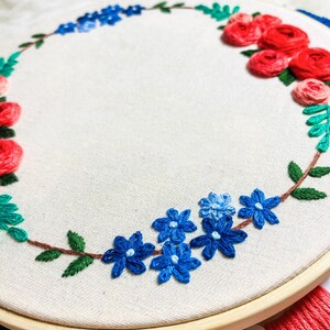 May include: A white embroidery hoop with a floral wreath design. The wreath is made of red, blue, and green embroidery floss. The flowers are arranged in a circle around the hoop.