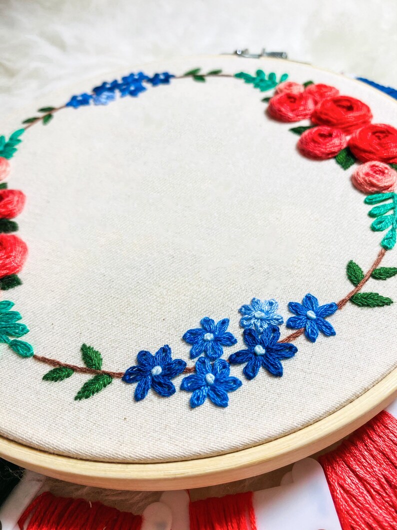 May include: A white embroidery hoop with a floral wreath design. The wreath is made of red, blue, and green embroidery floss. The flowers are arranged in a circle around the hoop.