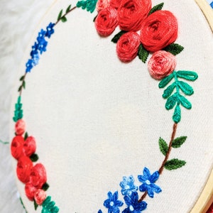 May include: A white embroidery hoop with a floral wreath design. The wreath is made of red, blue, and green embroidery floss. The flowers are arranged in a circle around the hoop.