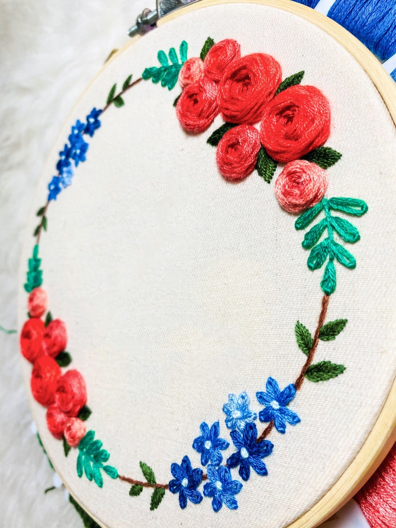 May include: A white embroidery hoop with a floral wreath design. The wreath is made of red, blue, and green embroidery floss. The flowers are arranged in a circle around the hoop.