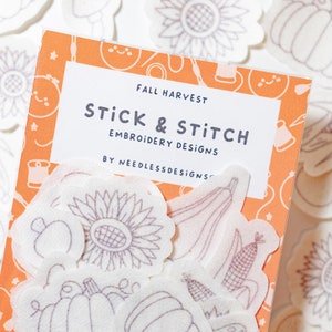 May include: A set of white felt embroidery designs with an orange background. The designs include a sunflower, pumpkin, corn, and squash. The text on the package reads "Fall Harvest Stick & Stitch Embroidery Designs by Needless Designs".