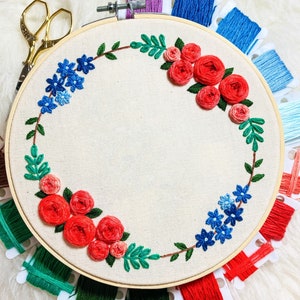 May include: A wooden embroidery hoop with a white fabric stretched across it. The fabric is embroidered with a wreath of red roses and blue flowers. The hoop is surrounded by spools of embroidery floss in various colors.
