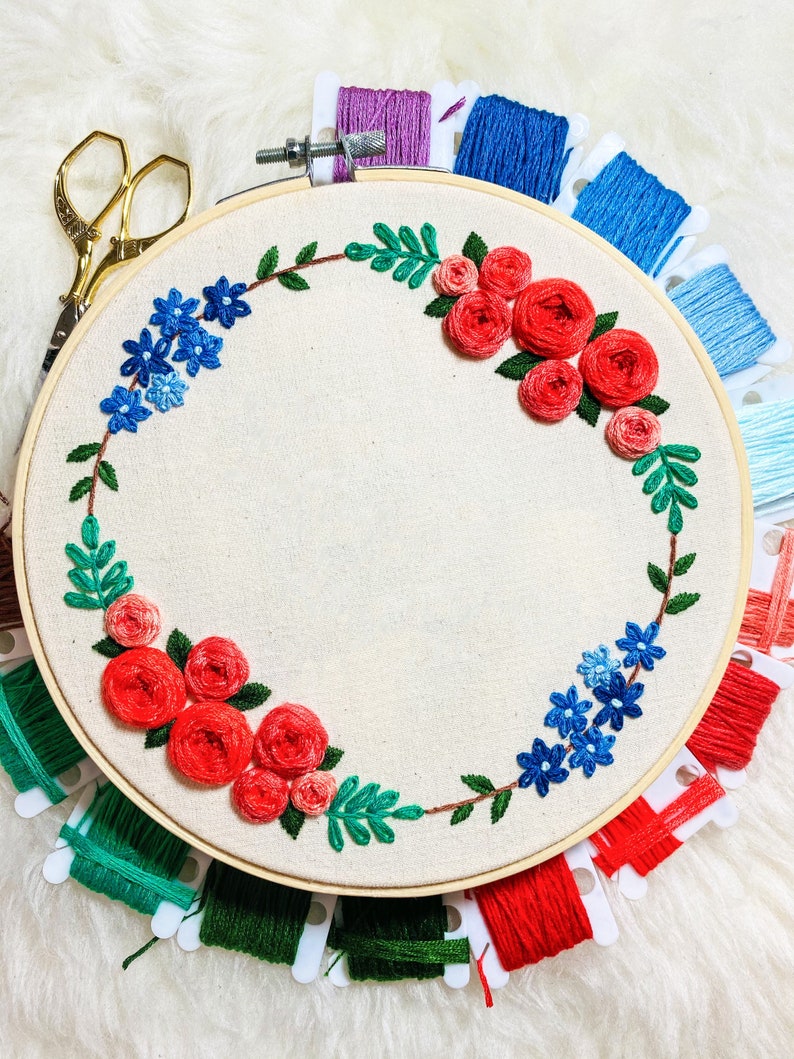 May include: A wooden embroidery hoop with a white fabric stretched across it. The fabric is embroidered with a wreath of red roses and blue flowers. The hoop is surrounded by spools of embroidery floss in various colors.