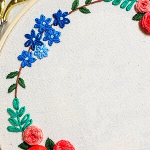 May include: Embroidered hoop art featuring a wreath of red roses, blue flowers, and green leaves on a white fabric background.