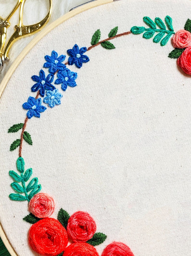 May include: Embroidered hoop art featuring a wreath of red roses, blue flowers, and green leaves on a white fabric background.