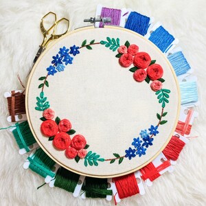 May include: A round embroidery hoop with a floral design featuring red roses and blue flowers. The hoop is surrounded by spools of embroidery floss in various colors, including red, green, blue, and pink.