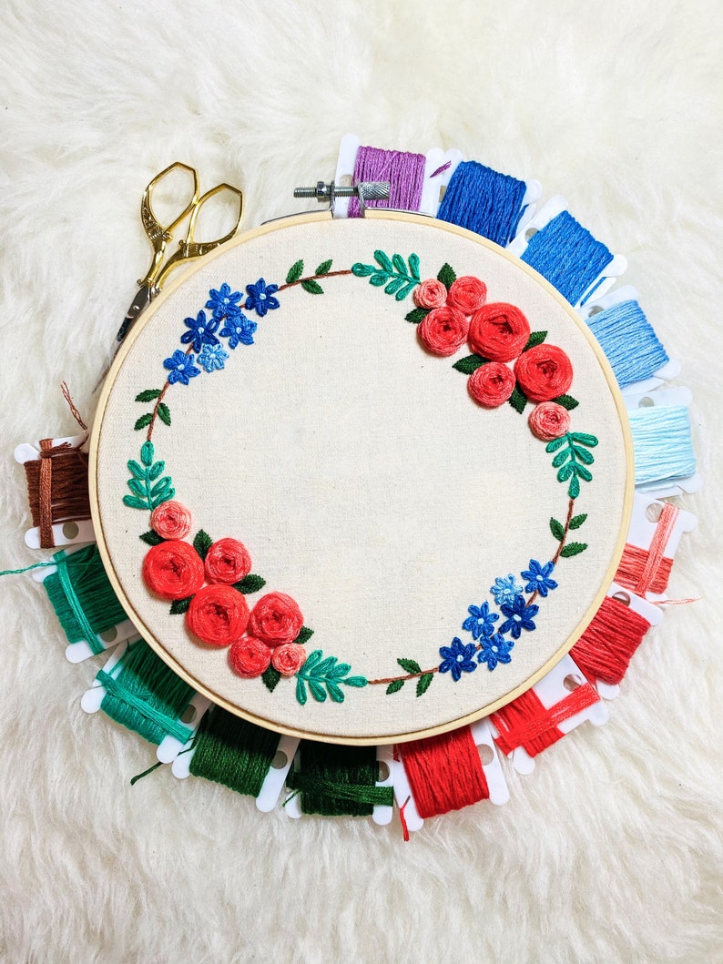 May include: A round embroidery hoop with a floral design featuring red roses and blue flowers. The hoop is surrounded by spools of embroidery floss in various colors, including red, green, blue, and pink.