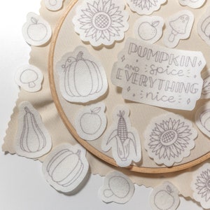 May include: A collection of iron-on patches featuring fall-themed designs, including pumpkins, sunflowers, corn, and an apple. The patches are arranged on a beige fabric background with the text "Pumpkin and spice everything nice" in a white banner.