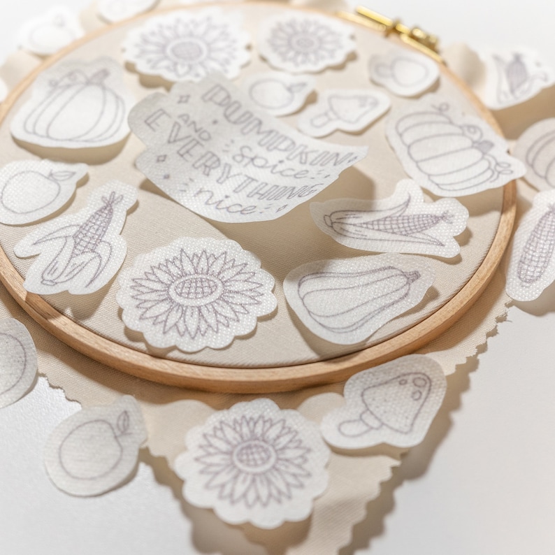 May include: A white embroidery hoop with various embroidered designs, including pumpkins, sunflowers, corn, and apples. The hoop is on a beige fabric background. A white piece of fabric with the text "Pumpkin spice and every thing nice!" is on top of the hoop.